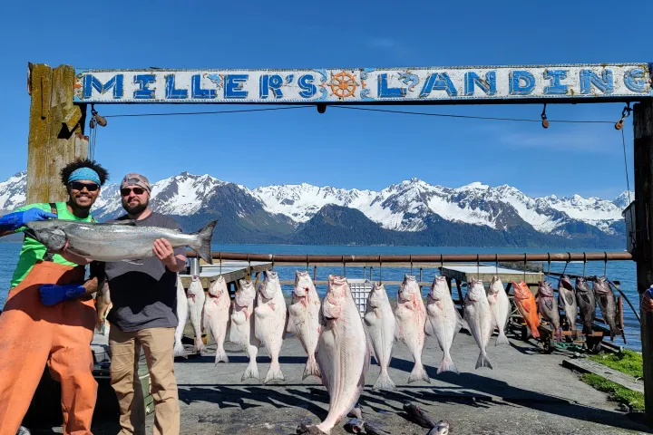 Two people holding a large fish in front of a row of hanging fish with snowy mountains in the background.