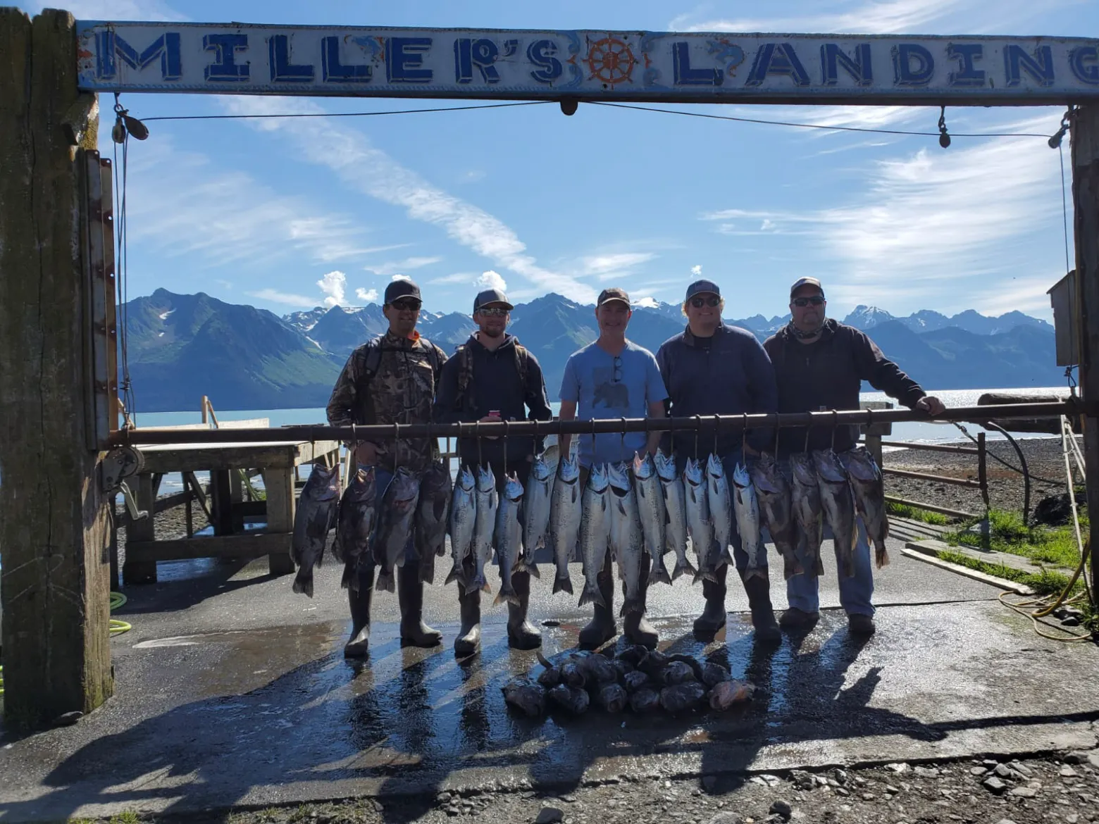 Five people standing with a rack of fish under a sign at a scenic lakeside location.