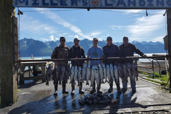 Five people standing with a rack of fish under a sign at a scenic lakeside location.