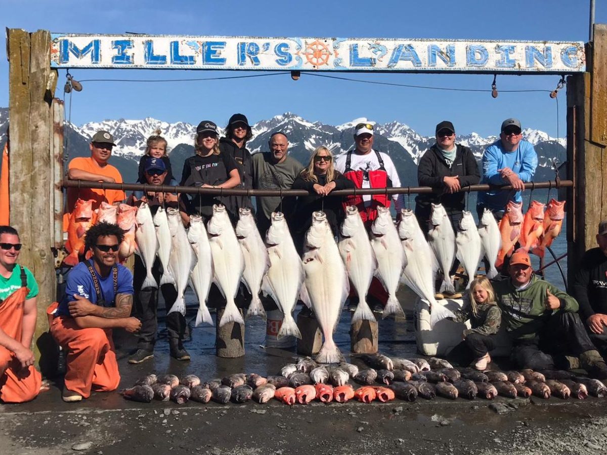 Group posing with large fish catch at Miller's Landing, snowy mountains in background.