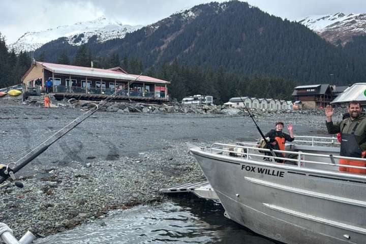 Two people on a boat near a rocky shore with a building, trees, and snowy mountains in the background.