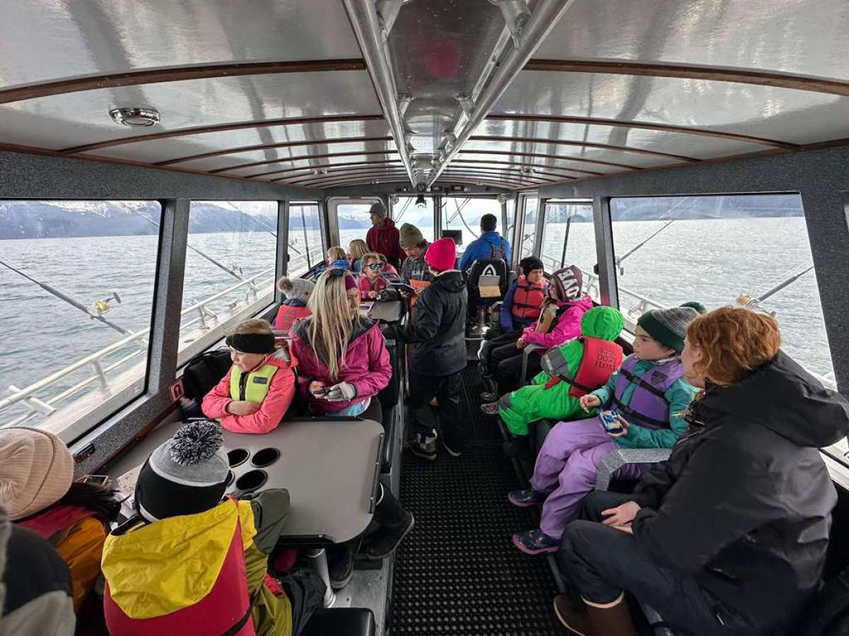 Passengers, including children, sitting inside a boat with large windows overlooking the sea.