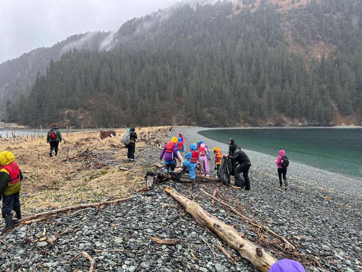 People in colorful coats walk on a rocky beach near a forested mountain.