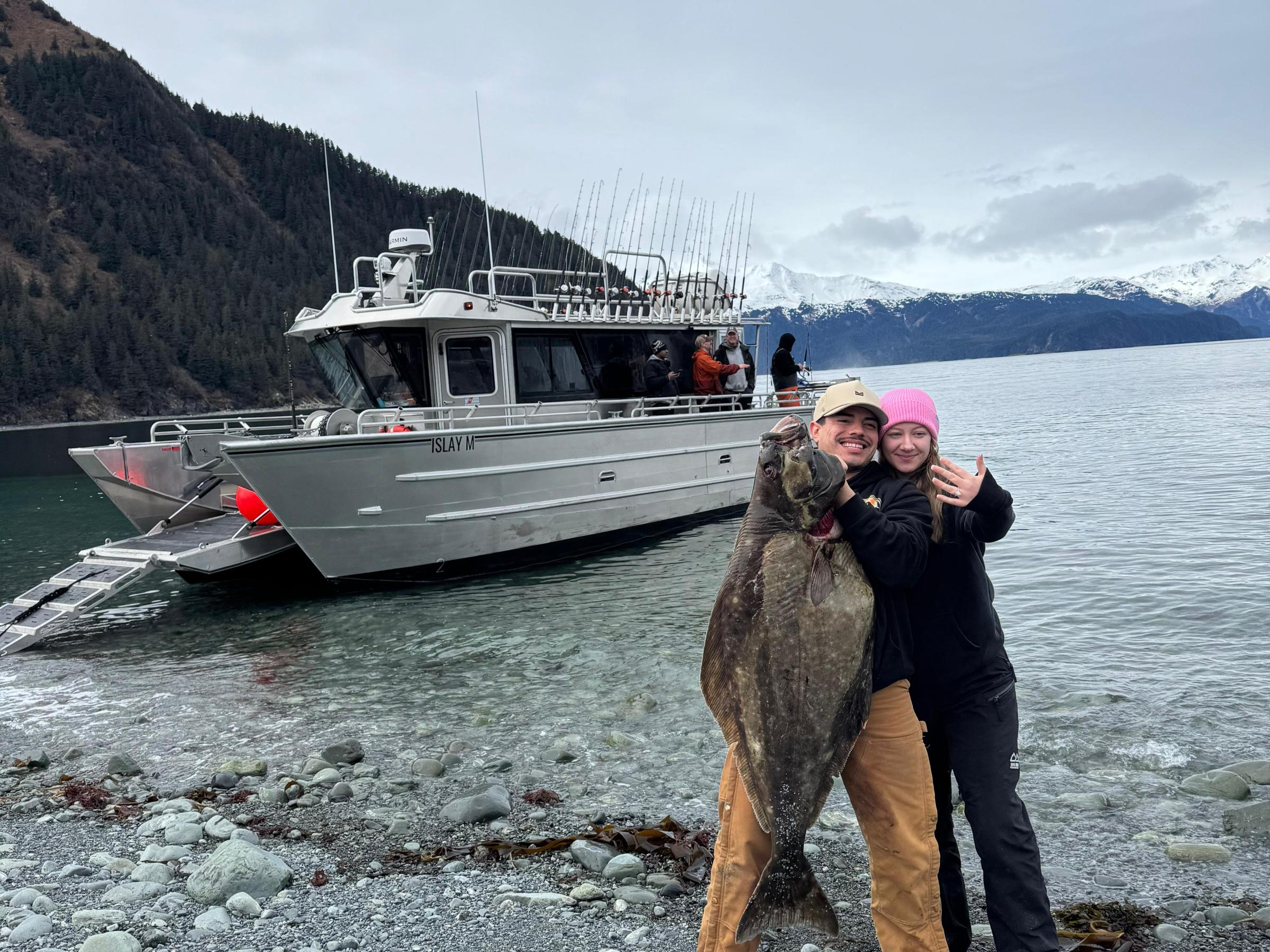 Two people on a rocky shore holding a large fish, with a boat and mountains in the background.