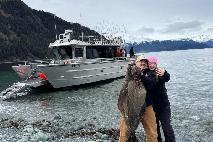Two people on a rocky shore holding a large fish, with a boat and mountains in the background.