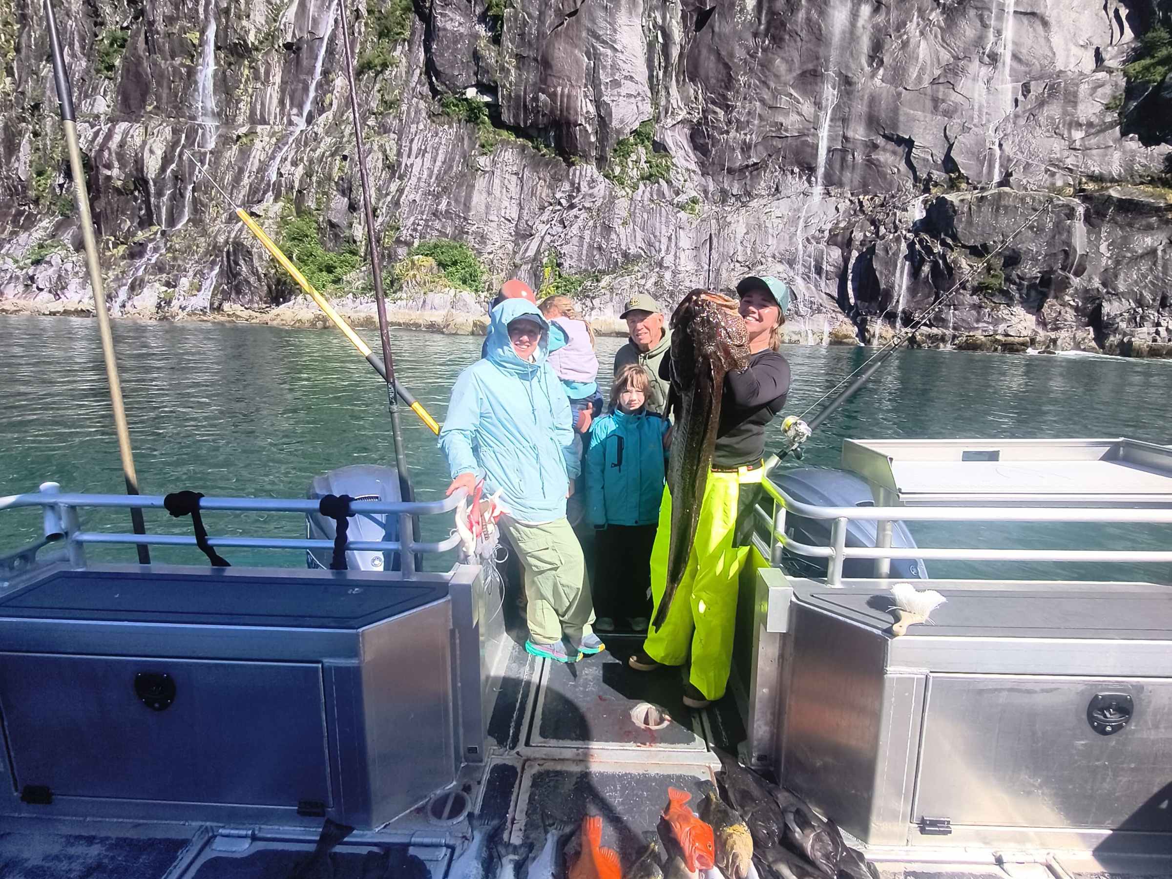 Group on boat with large fish catch, rocky cliff in background.