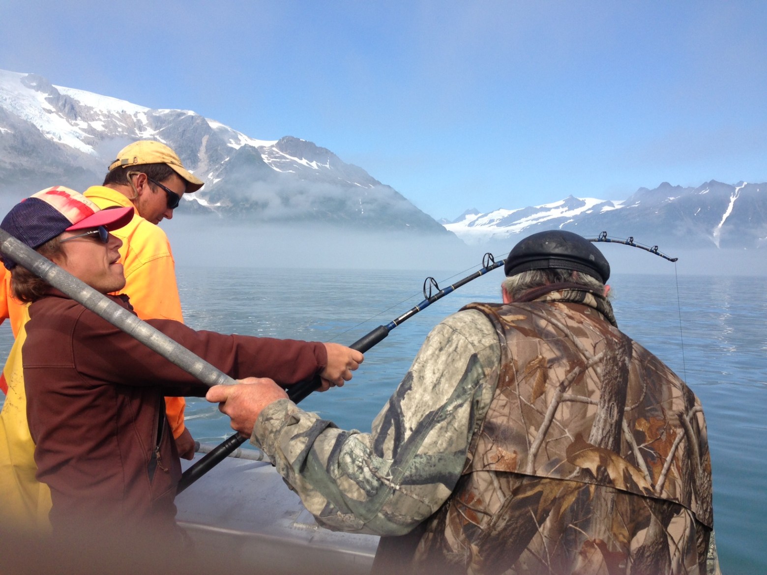 Three people fishing on a boat with snowy mountains in the background.