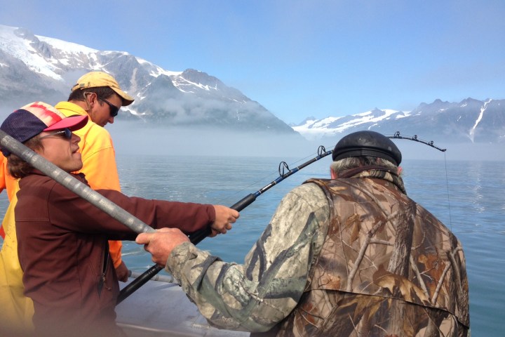 Three people fishing on a boat with snowy mountains in the background.