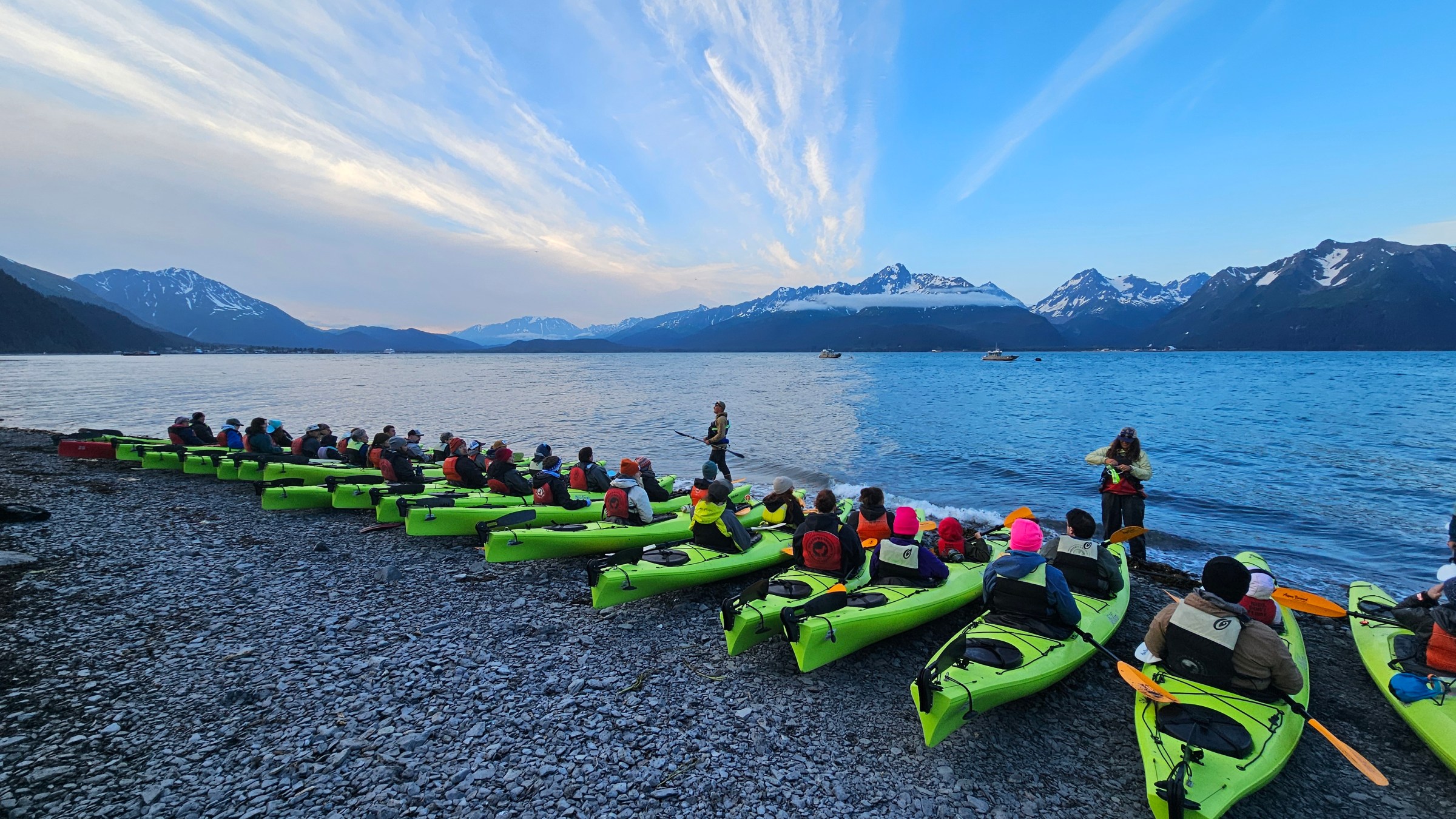 Group of people in kayaks on a rocky beach with mountains in the background at sunset.