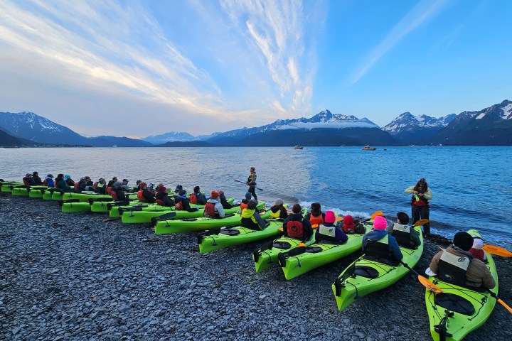 Group of people in kayaks on a rocky beach with mountains in the background at sunset.
