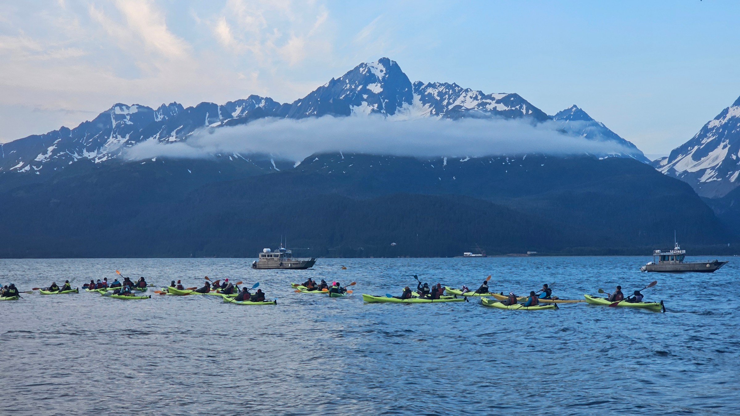 Group of people kayaking on a lake with snow-capped mountains in the background under a cloudy sky.
