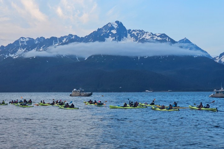 Group of people kayaking on a lake with snow-capped mountains in the background under a cloudy sky.