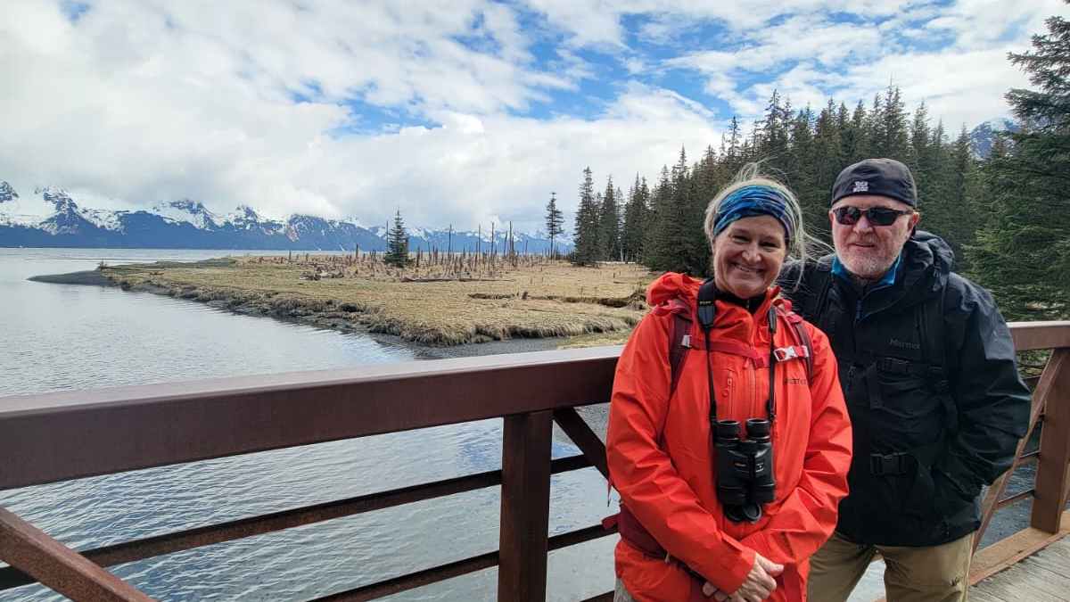 Two people in outdoor clothing on a bridge with mountains and trees in the background.