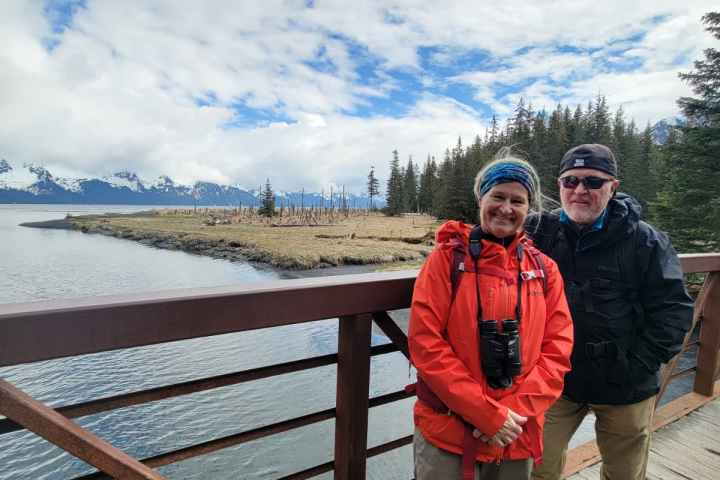 Two people in outdoor clothing on a bridge with mountains and trees in the background.
