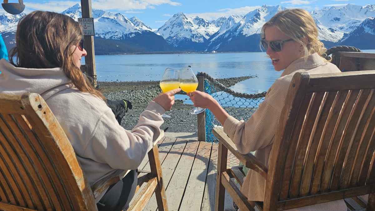 Two people toast with drinks on a deck overlooking snowy mountains and a lake.