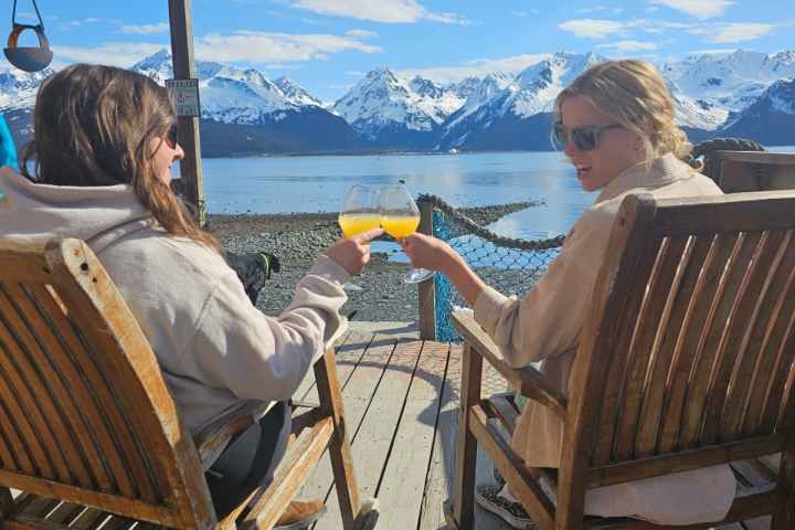 Two people toast with drinks on a deck overlooking snowy mountains and a lake.