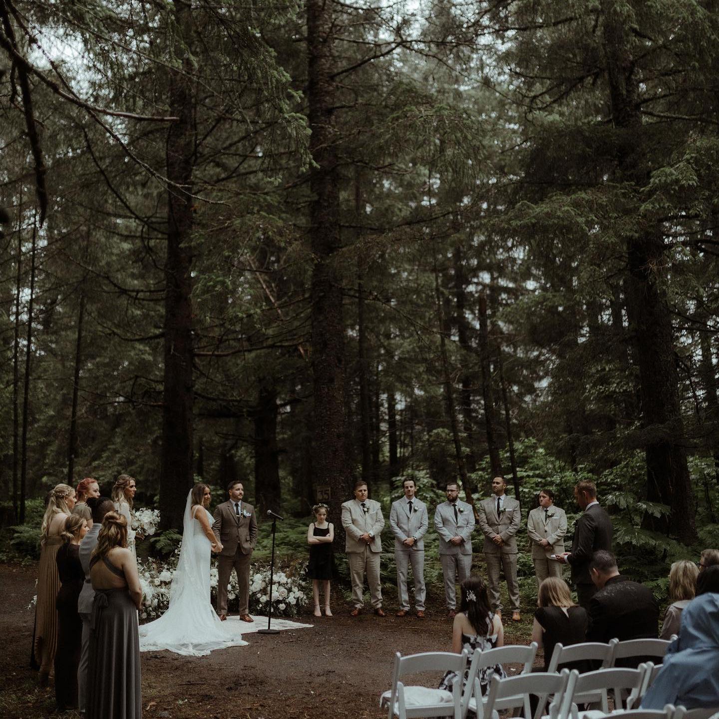 Outdoor wedding ceremony in a forest with a bridal party and guests seated on white chairs.