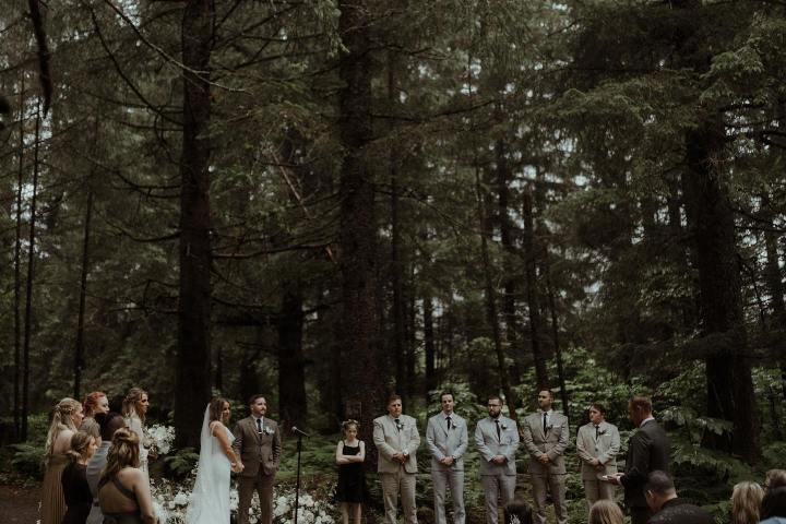 Outdoor wedding ceremony in a forest with a bridal party and guests seated on white chairs.