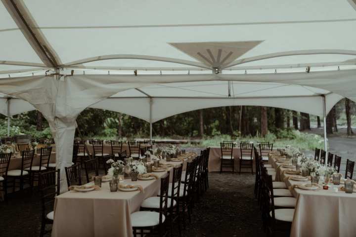 Outdoor wedding reception setup under a tent with rows of tables and wooden chairs.