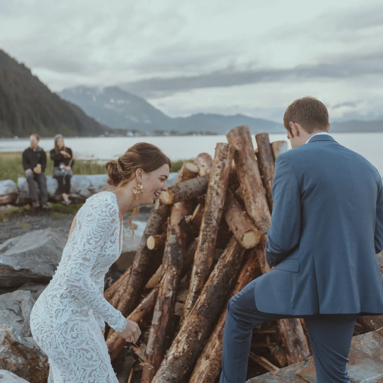 Bride and groom near a woodpile by a lake, with mountains in the background.