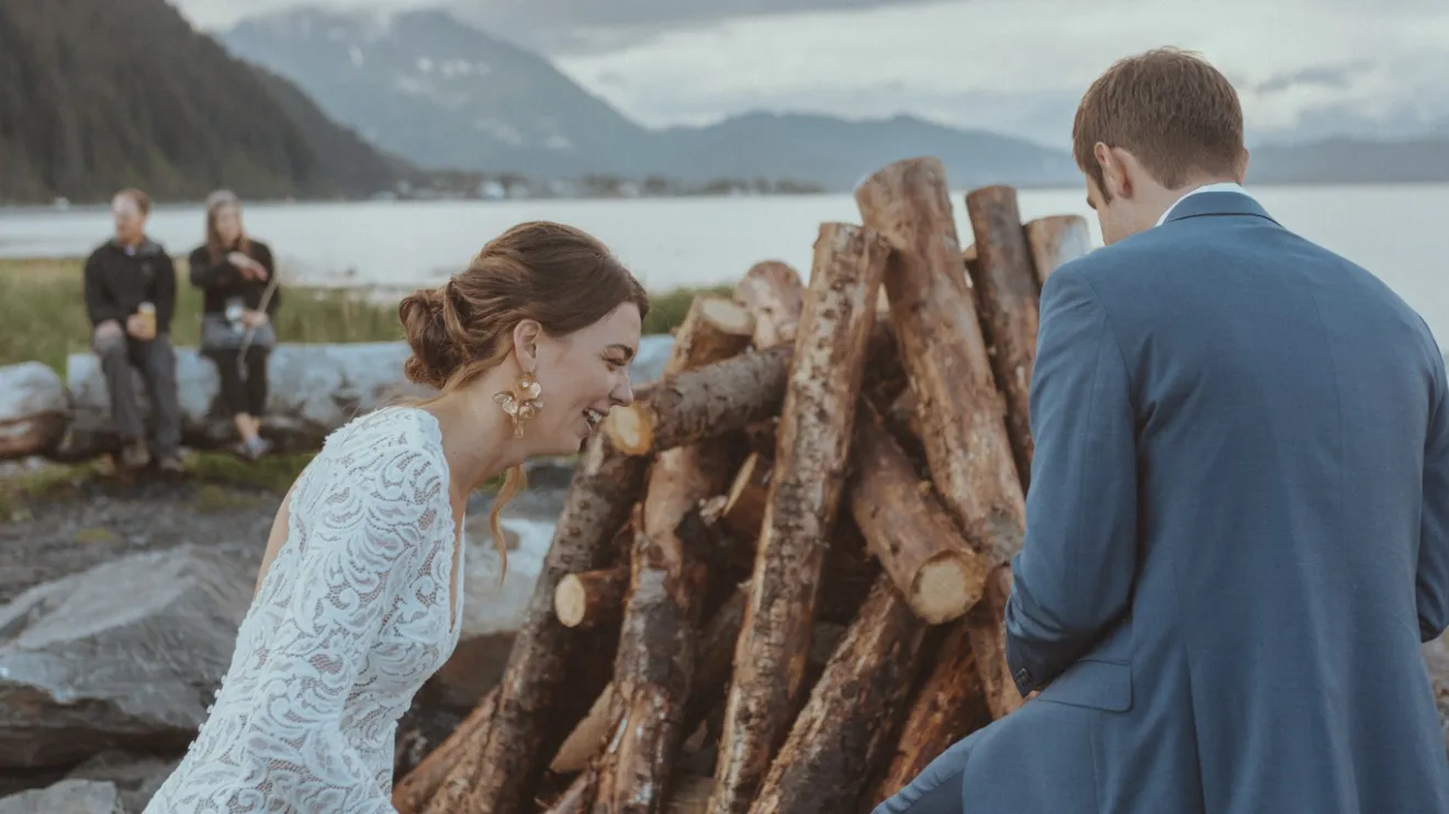 Bride and groom near a woodpile by a lake, with mountains in the background.
