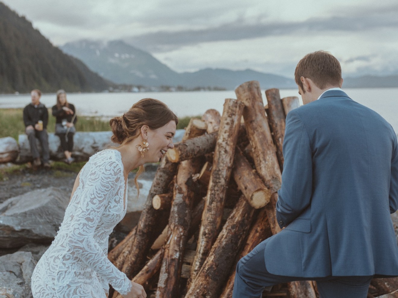 Bride and groom near a woodpile by a lake, with mountains in the background.
