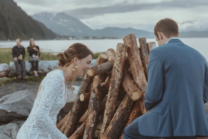 Bride and groom near a woodpile by a lake, with mountains in the background.