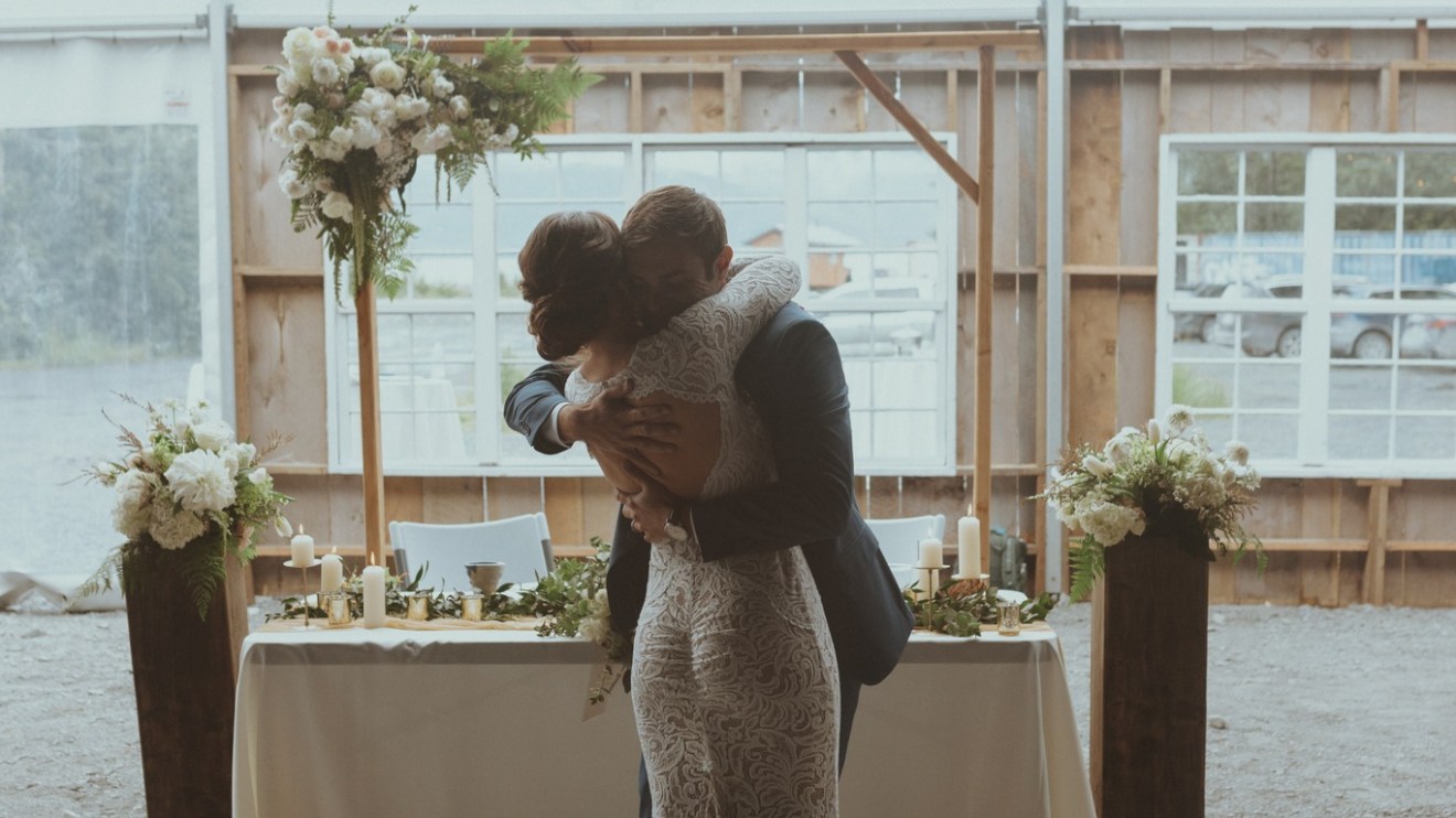 Couple hugging in front of a decorated table and flowers in a tented setting.