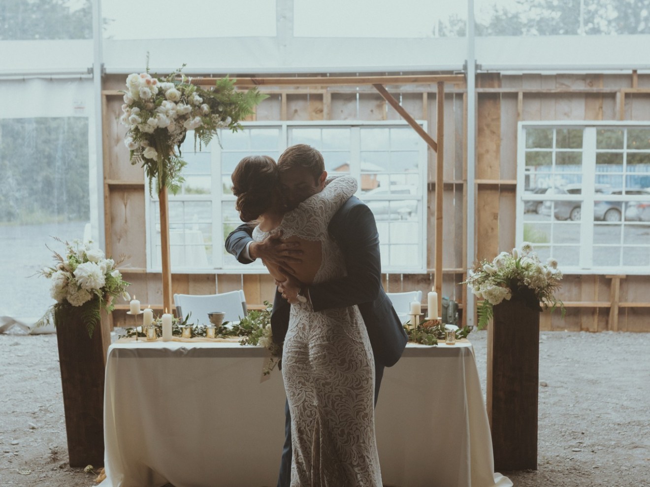 Couple hugging in front of a decorated table and flowers in a tented setting.