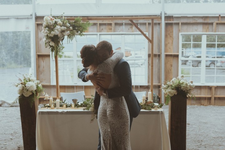 Couple hugging in front of a decorated table and flowers in a tented setting.