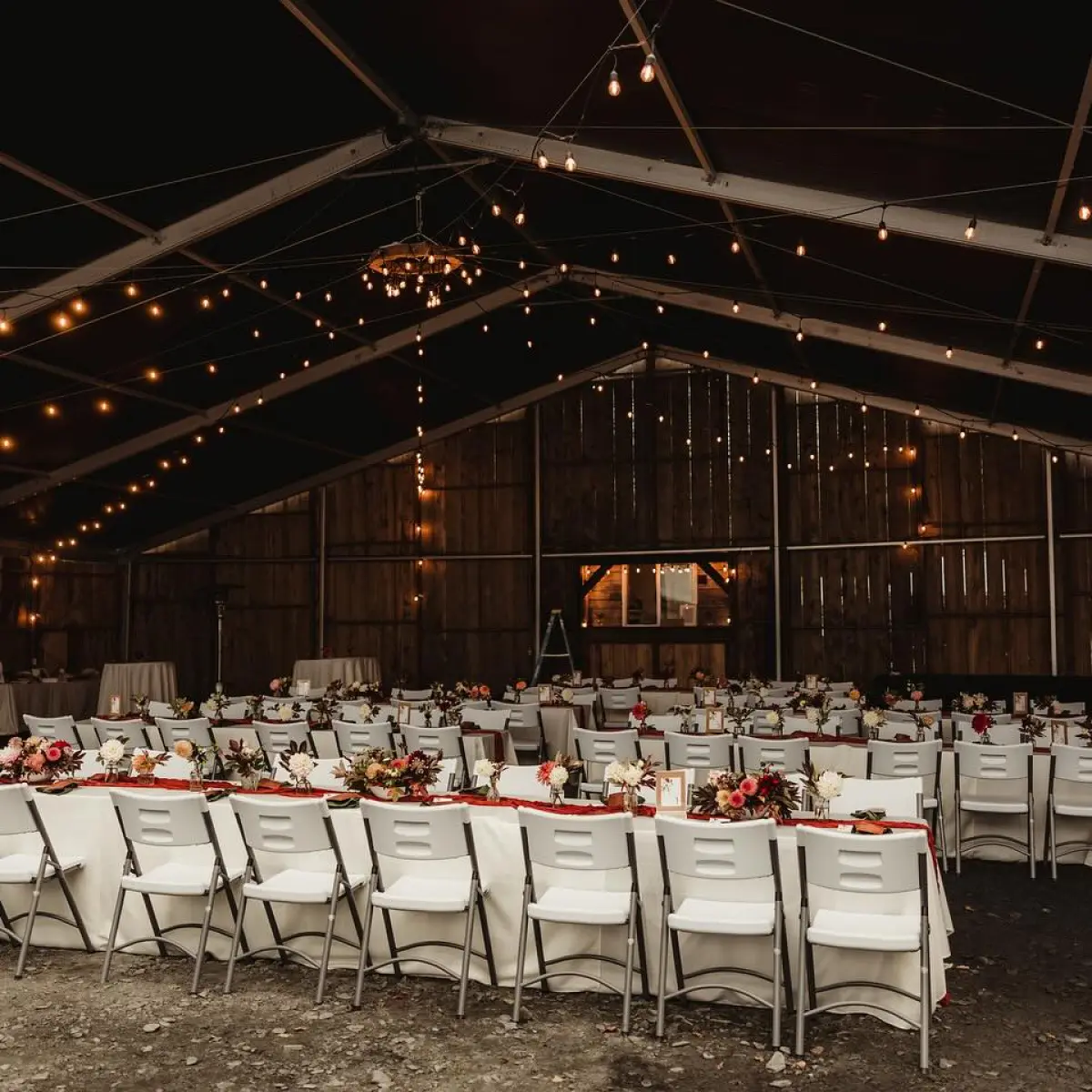 Wedding reception setup in a rustic barn with tables, chairs, floral centerpieces, and string lights.