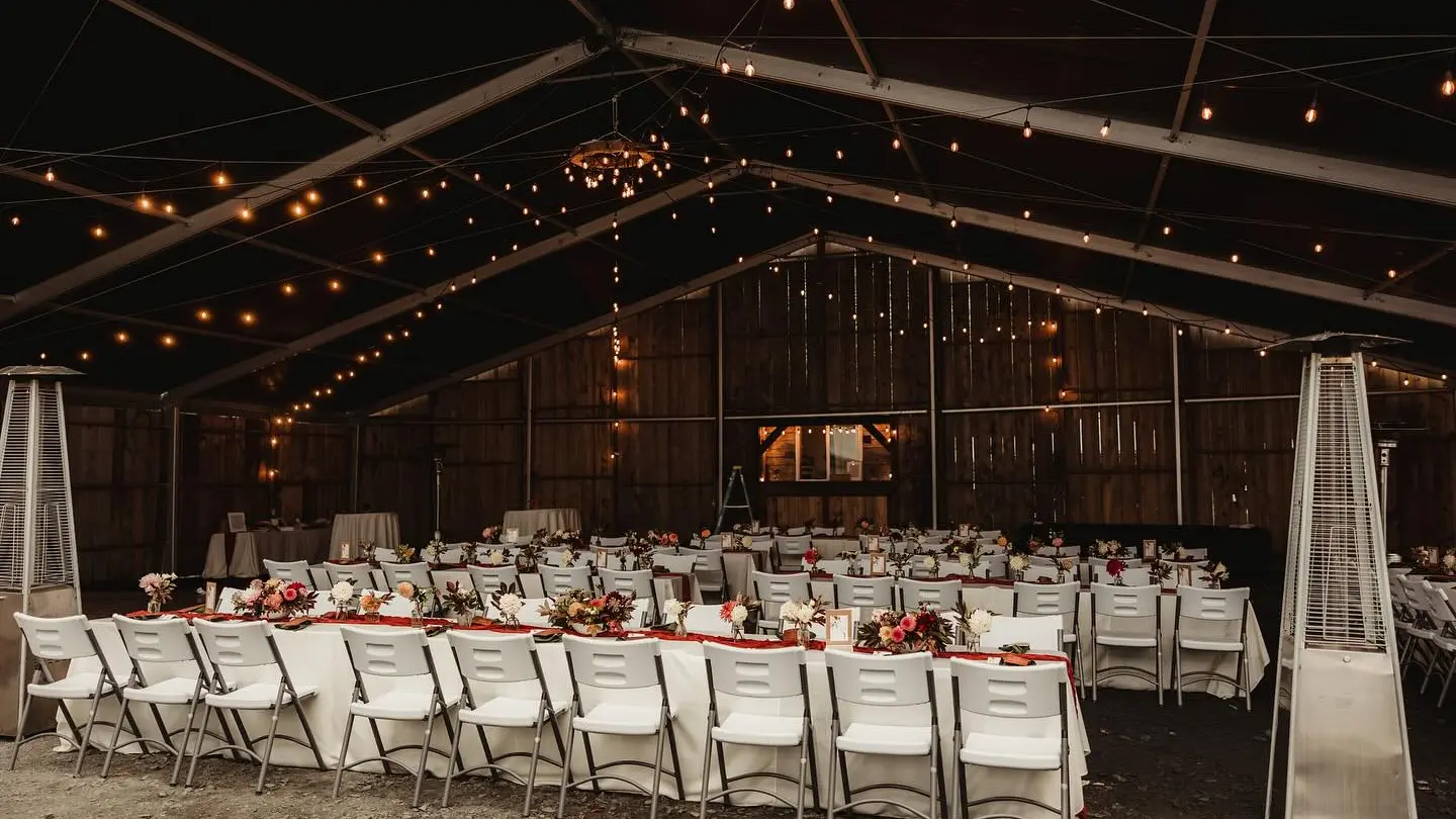 Wedding reception setup in a rustic barn with tables, chairs, floral centerpieces, and string lights.