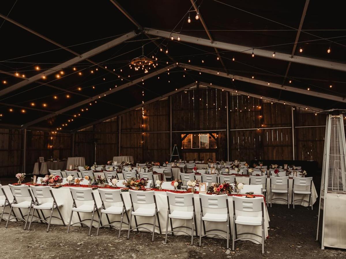 Wedding reception setup in a rustic barn with tables, chairs, floral centerpieces, and string lights.