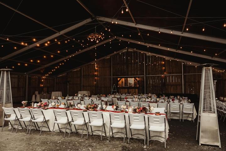 Wedding reception setup in a rustic barn with tables, chairs, floral centerpieces, and string lights.