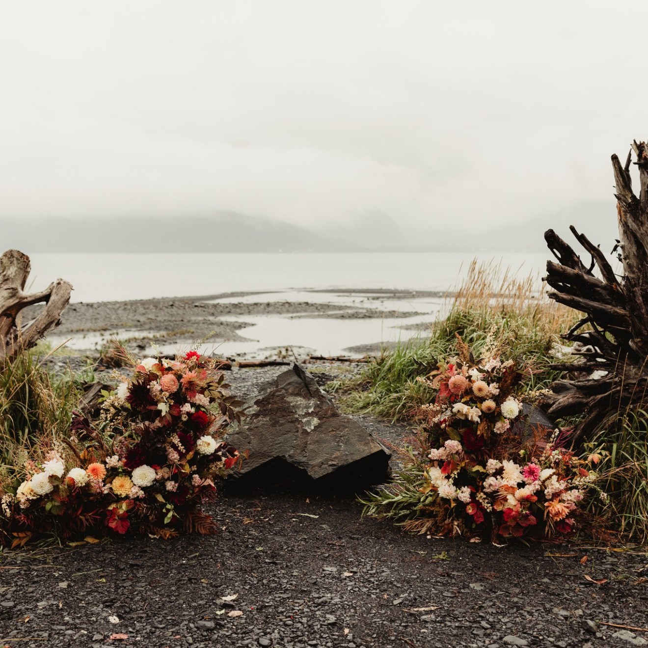Flower arrangements on beach with driftwood, gray sky, and mountains.