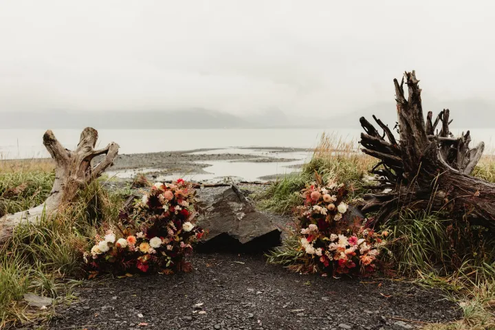 Flower arrangements on beach with driftwood, gray sky, and mountains.