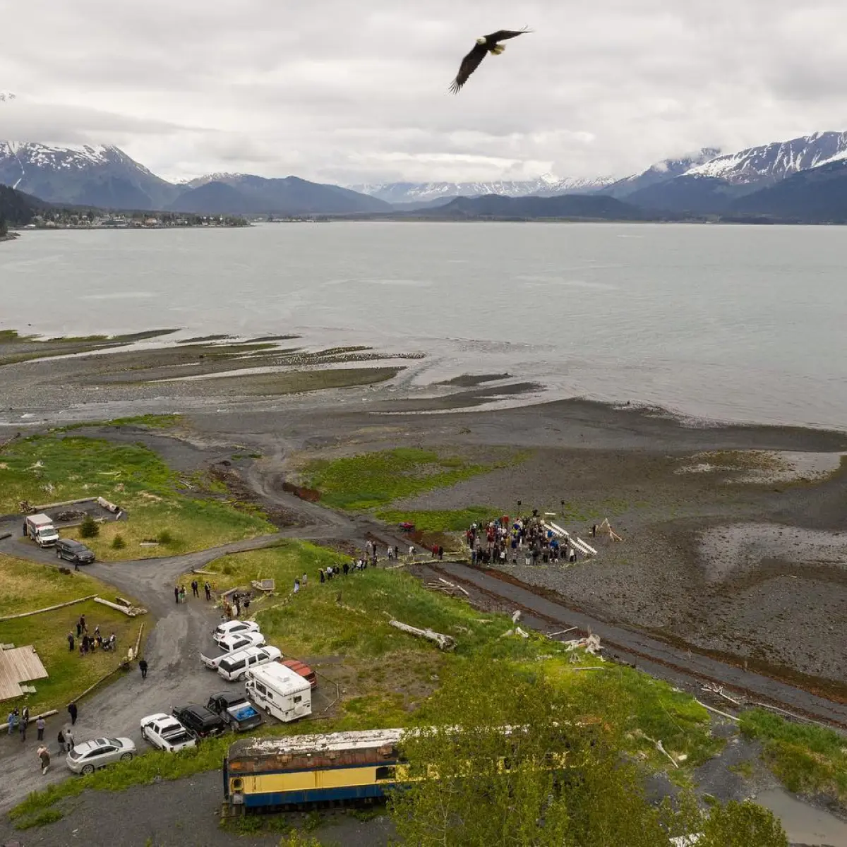 Aerial view of coastline with people gathered near shore, tents, and vehicles, mountains in the background.