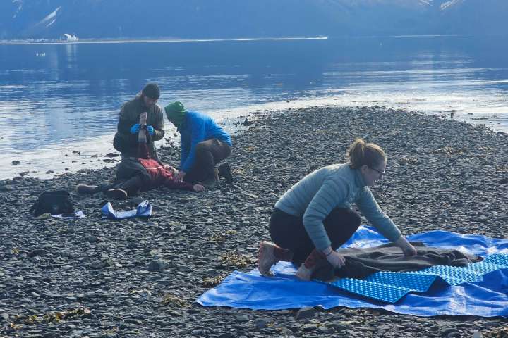 People on rocky beach giving first aid and setting up a tarp with mountains in background.