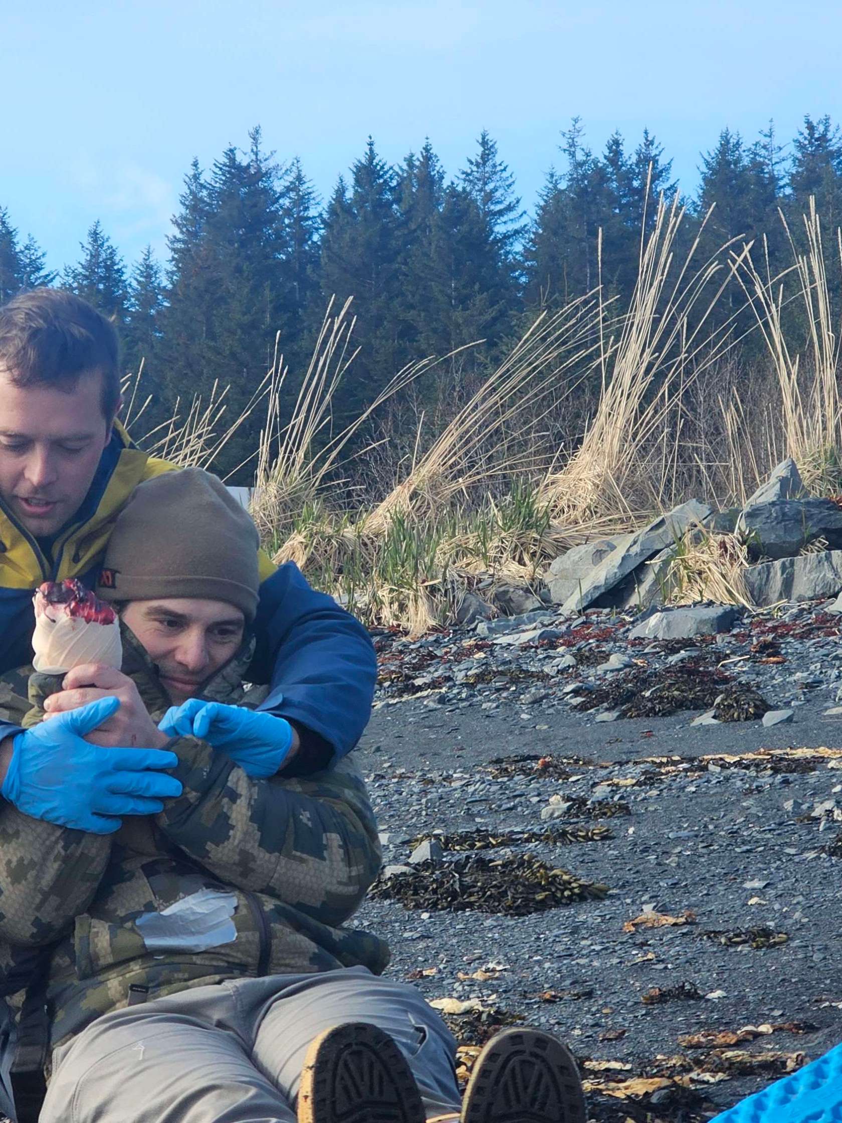 Three people on a rocky beach; two are sitting, and one is kneeling nearby with blue gear.