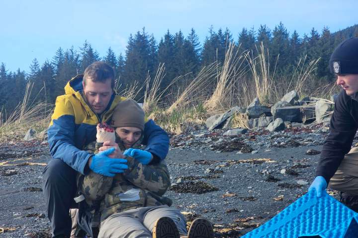 Three people on a rocky beach; two are sitting, and one is kneeling nearby with blue gear.