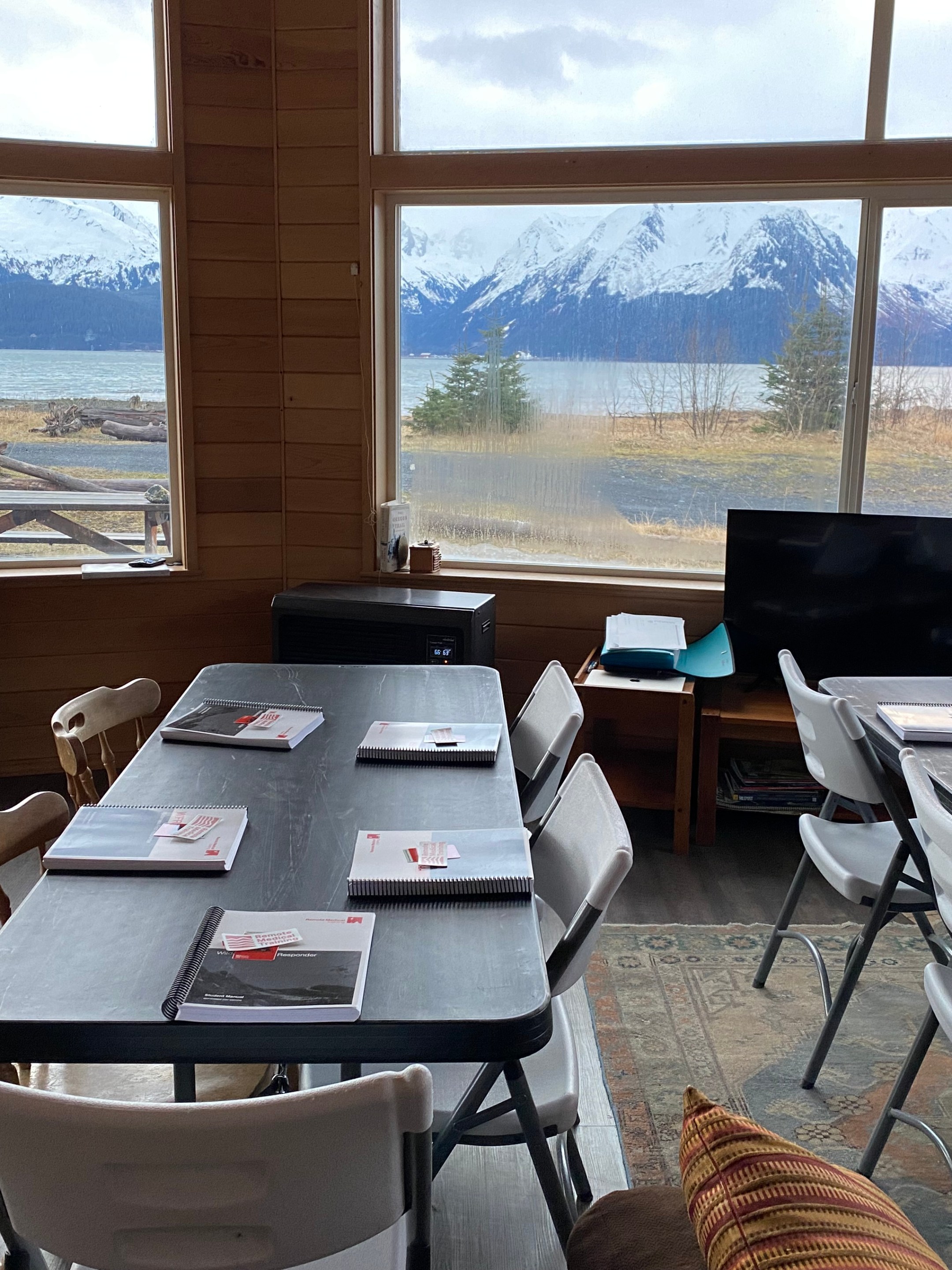 Room with tables, chairs, notebooks and large windows showing snowy mountains and a lake.