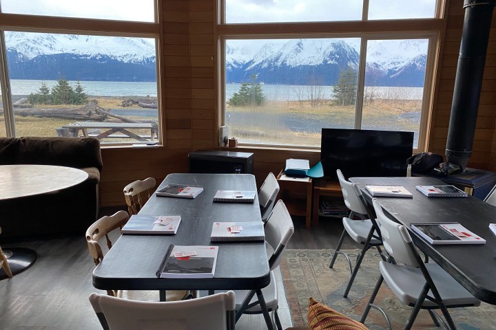 Room with tables, chairs, notebooks and large windows showing snowy mountains and a lake.