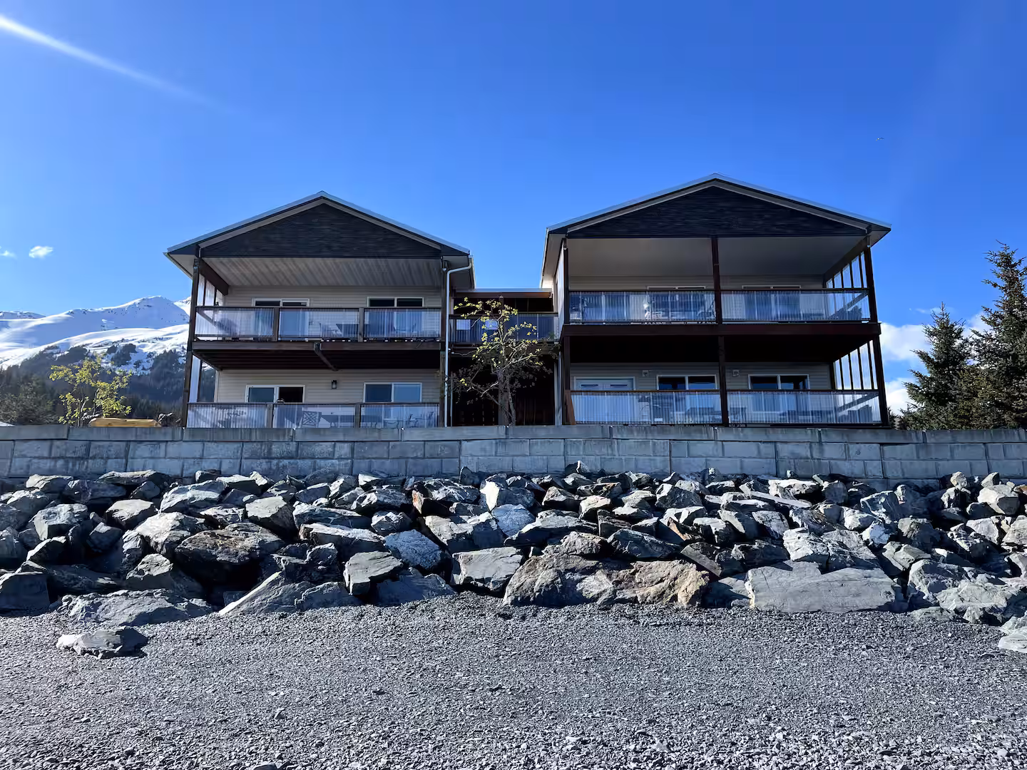 Two-story building with balconies, stone wall, and rocky foreground under clear blue sky.