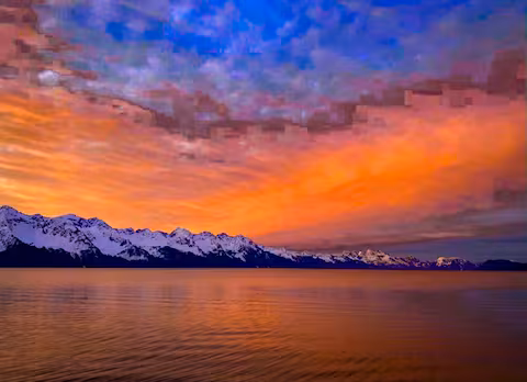 Snow-capped mountains at sunset with colorful sky and calm water reflection.