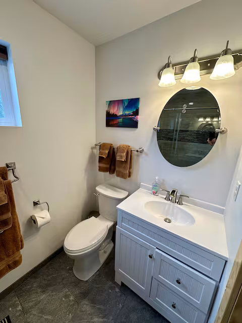 Small bathroom with round mirror, sink, toilet, towels, and landscape art on wall.