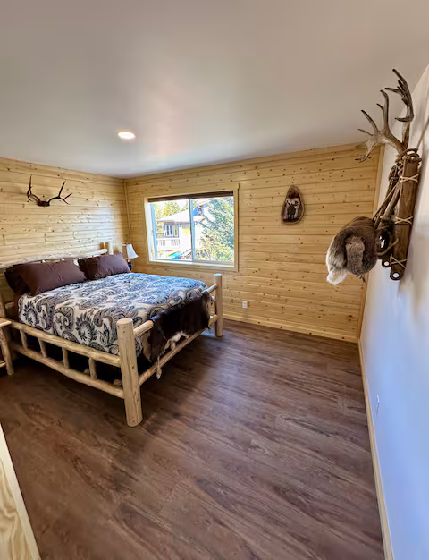 Rustic bedroom with wooden walls, antler decor, large window, and a bed with patterned bedding.