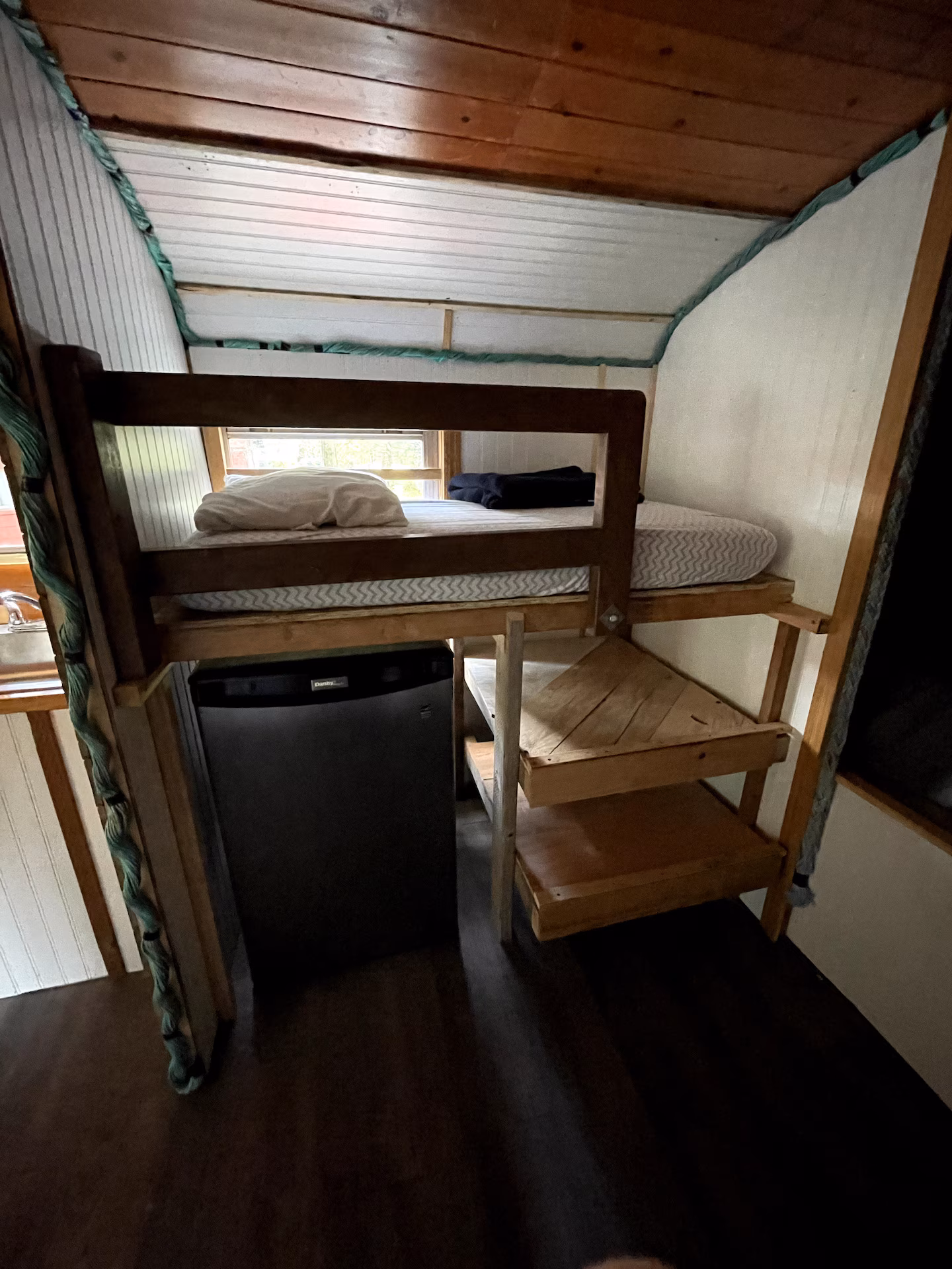Loft bed with ladder, fridge underneath, wooden ceiling.