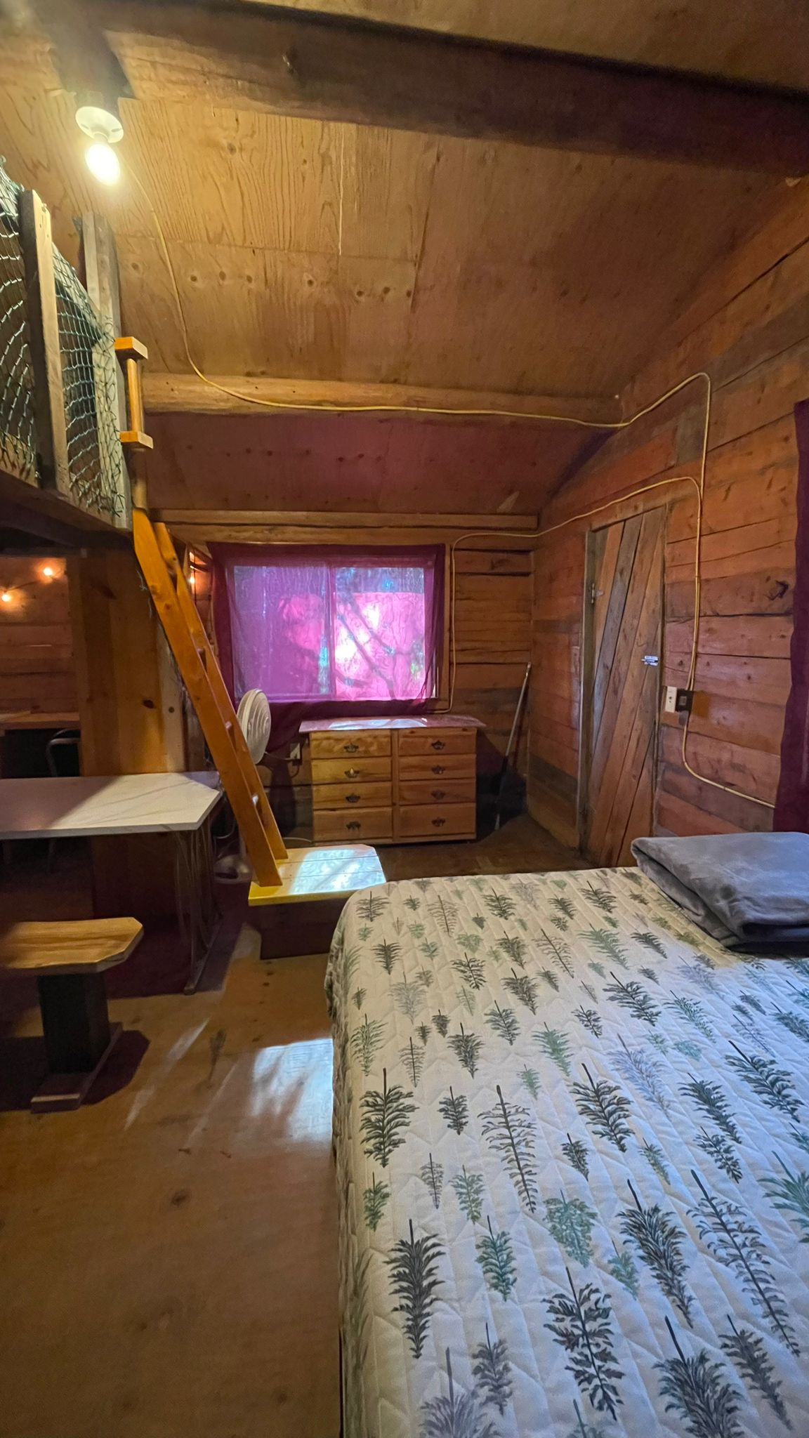 Cozy wooden cabin interior with bed, loft, dresser, desk, and string lights.