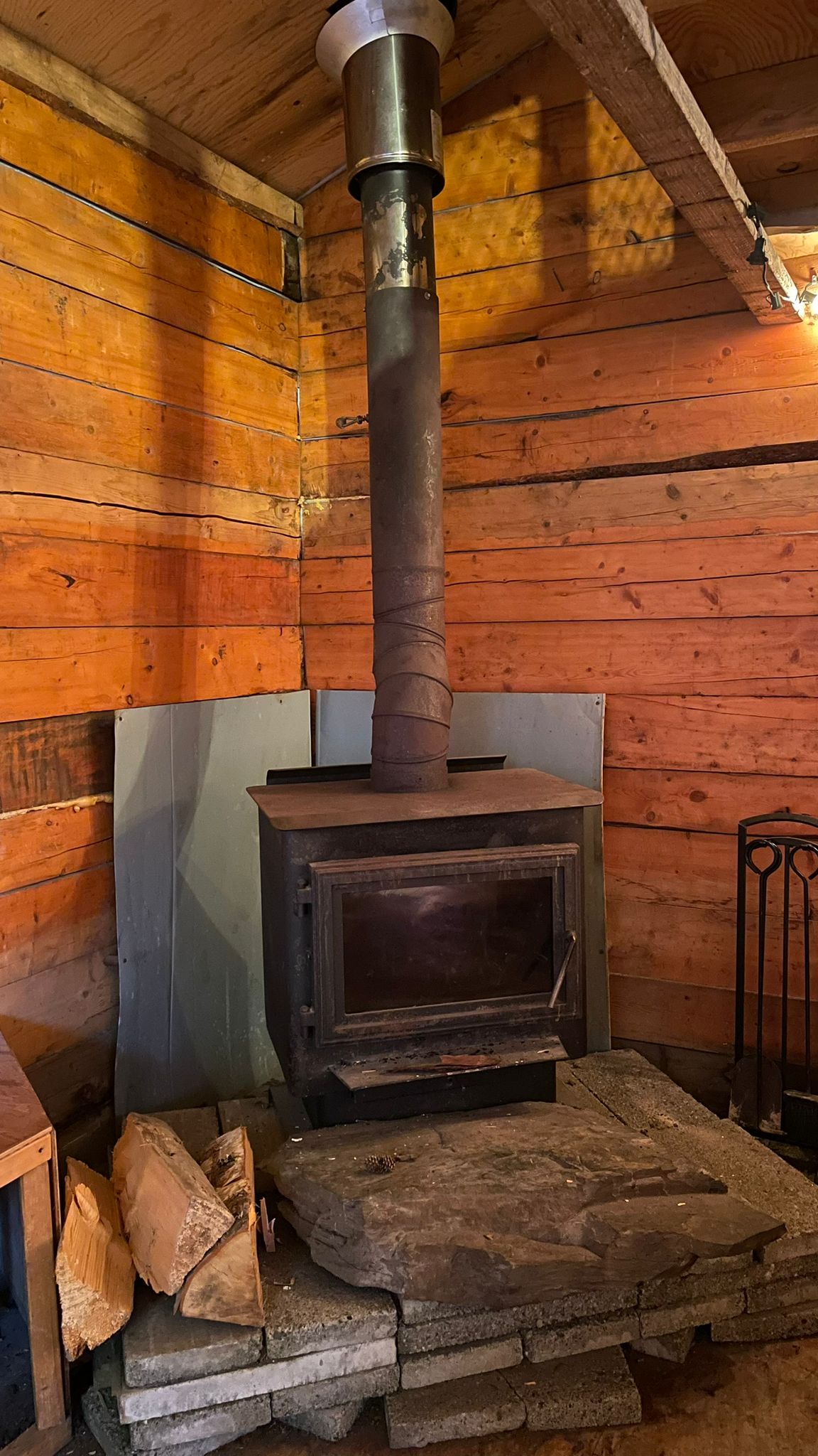 Wood stove with chimney in a rustic wooden room, logs stacked nearby.