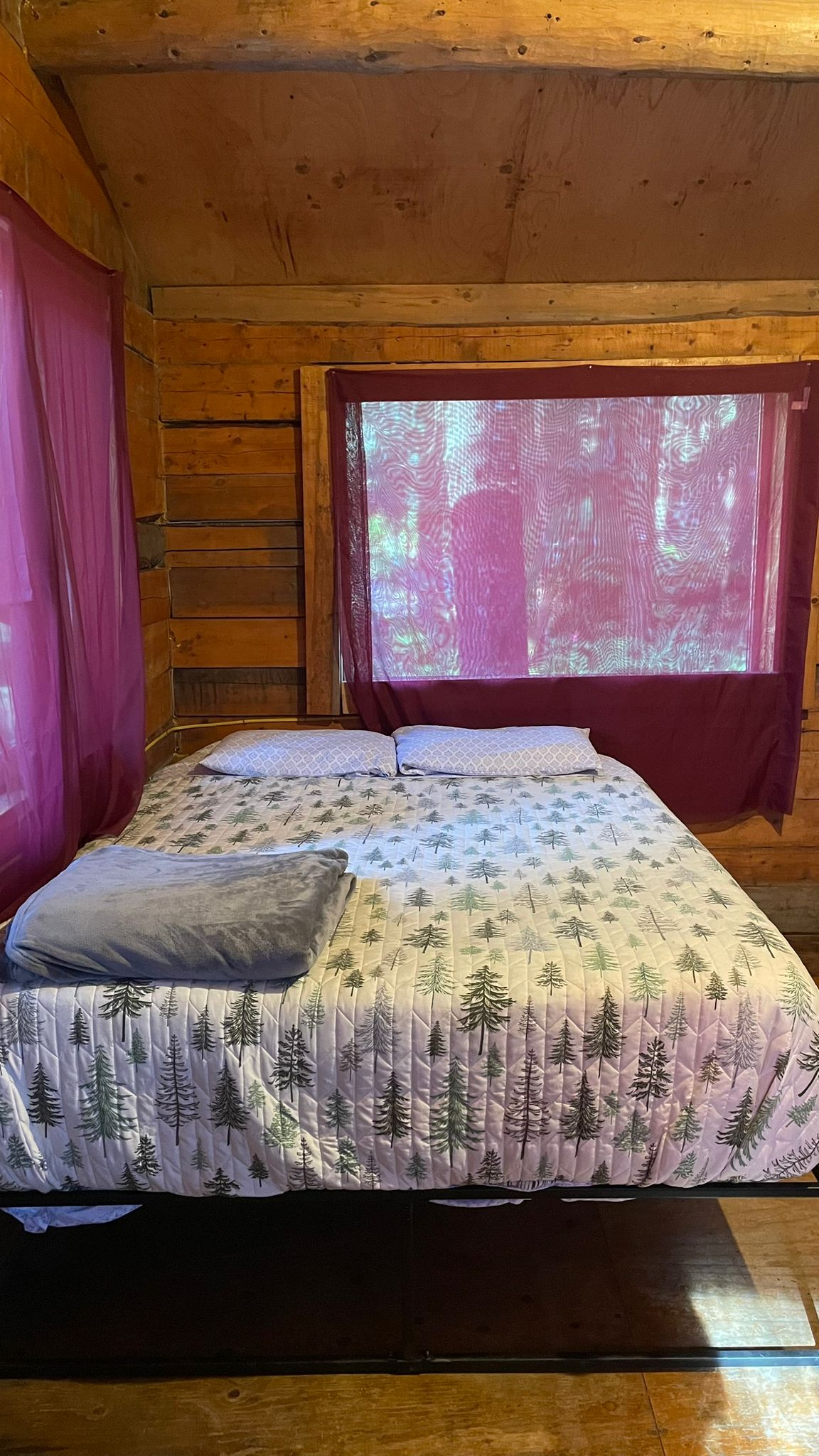 Cozy log cabin bedroom with bed and tree-patterned bedding beside purple curtained window.